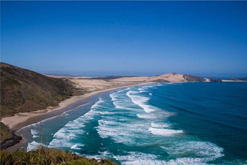 See the best places to go in New Zealand, view across sandy beach with foamy waves and a sandy peninsula in the distance under an azure blue sky
