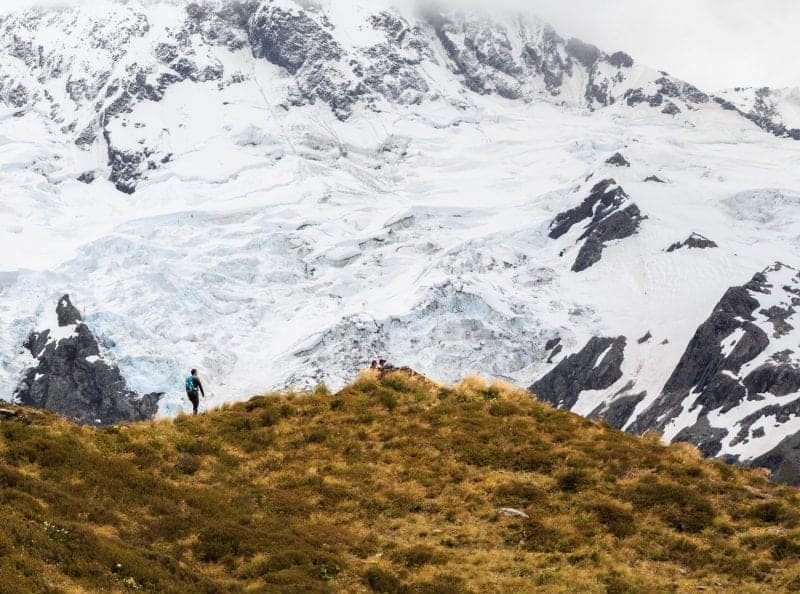 Discover the most beautiful places in New Zealand, man standing on grassy hilltop in front of huge snow-covered rocky mountain